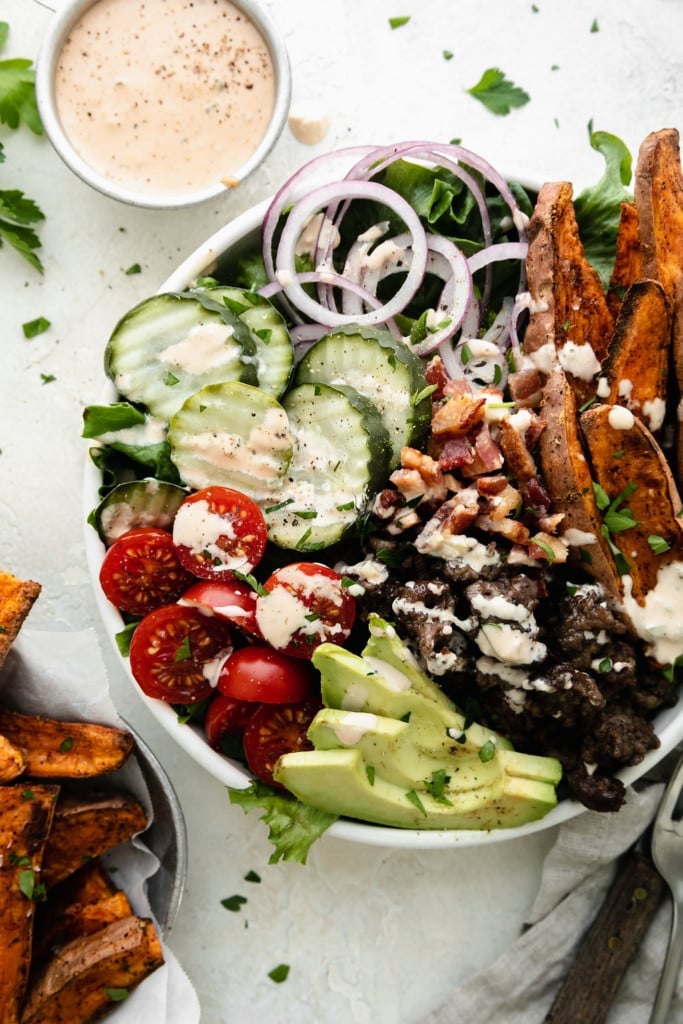 Overhead view of a burger bowl in a white bowl filled with mixed greens, hamburger, pickles, sweet potato fries, and drizzled with special sauce. 