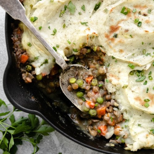 Overhead view of vegan shepherd's pie in a cast iron skillet with a spoon scooping up a serving of lentil and gravy filling.