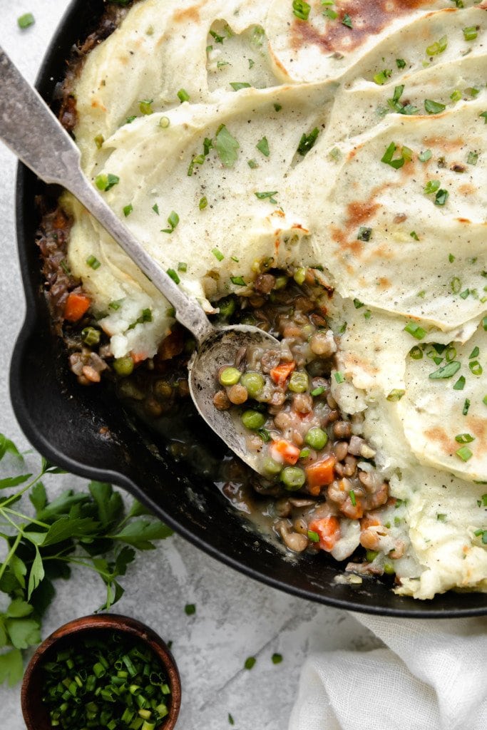 Overhead view of vegan shepherd's pie in a cast iron skillet with a spoon scooping up a serving of lentil and gravy filling.