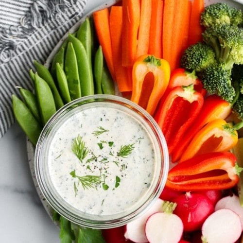 Overhead view of creamy Paleo ranch dressing in a short glass jar on a platter of brightly colored freshly cut vegetables.