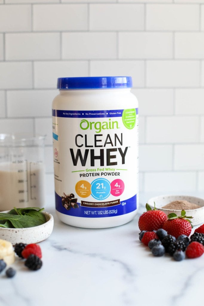 A container of Orgain Clean Whey protein powder against a white background with fresh fruit in the forefront. 