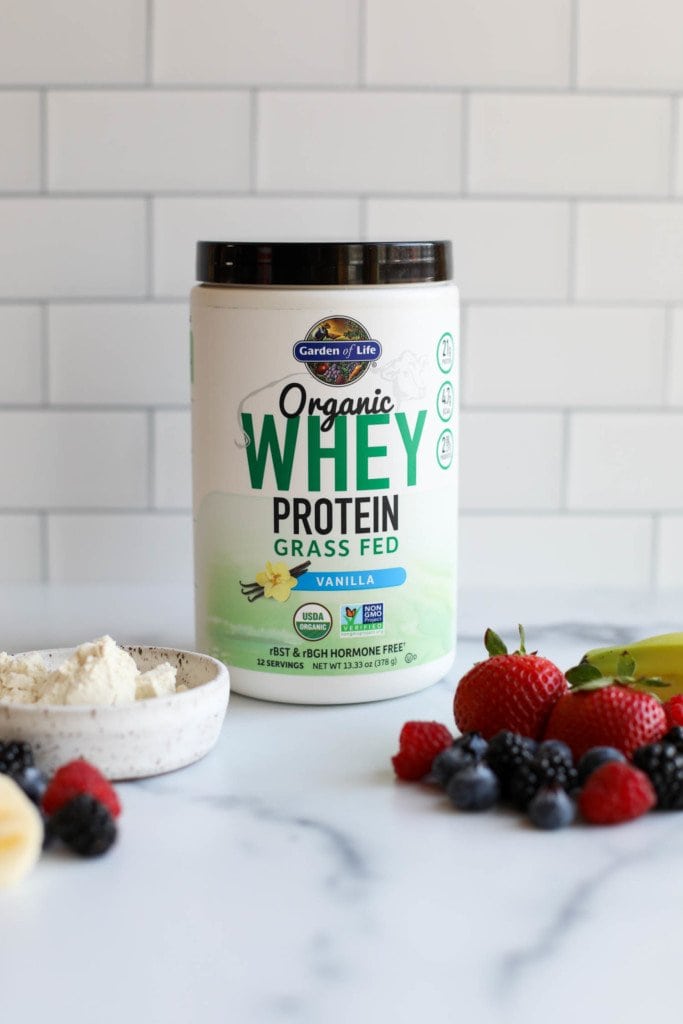 A container of Garden of Life Organic Whey Protein powder against a white background with fresh fruit in the forefront. 