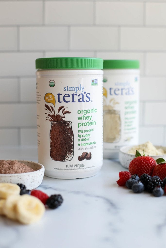 Two containers of Simply Tera's organic whey protein powder against a white background with fresh fruit in the forefront. 