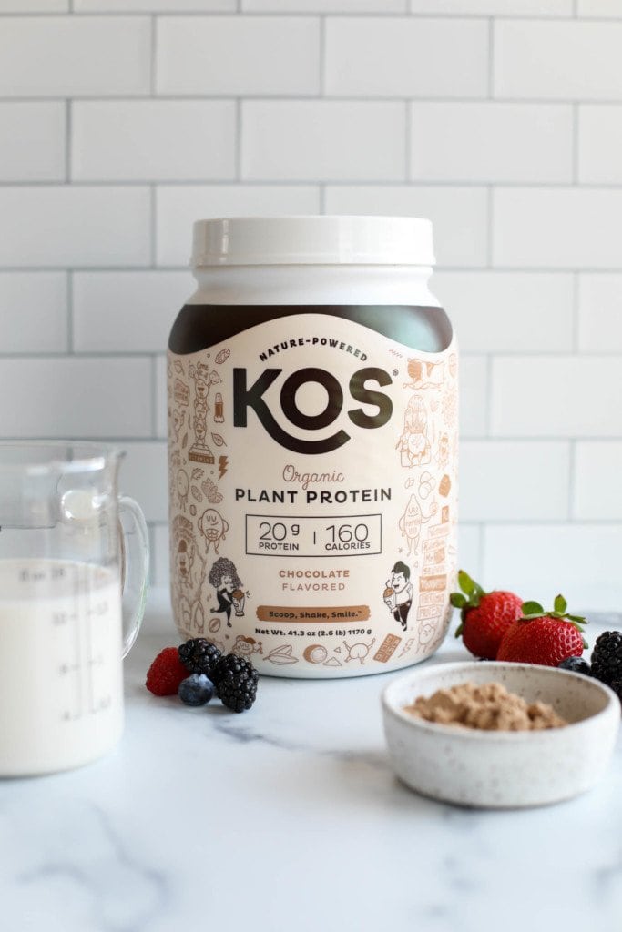 A container of KOS plant based protein powder against a white background, with berries and protein powder in a small bowl in the forefront. 