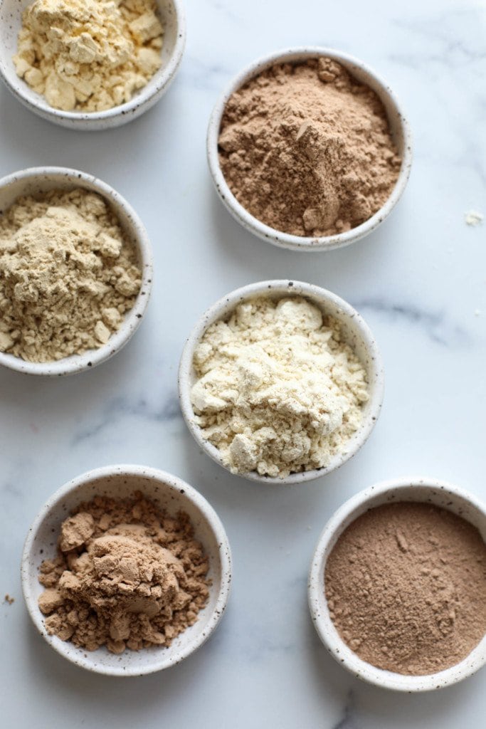 Overhead view of several protein powders in small bowls arranged together on a marble countertop. 