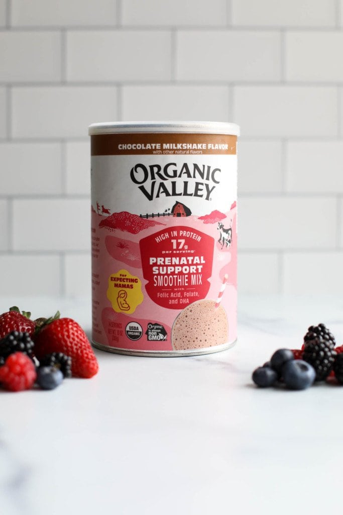 A container of Organic Valley prenatl support smoothie mix whey protein powder against a white background with fresh fruit in the forefront. 