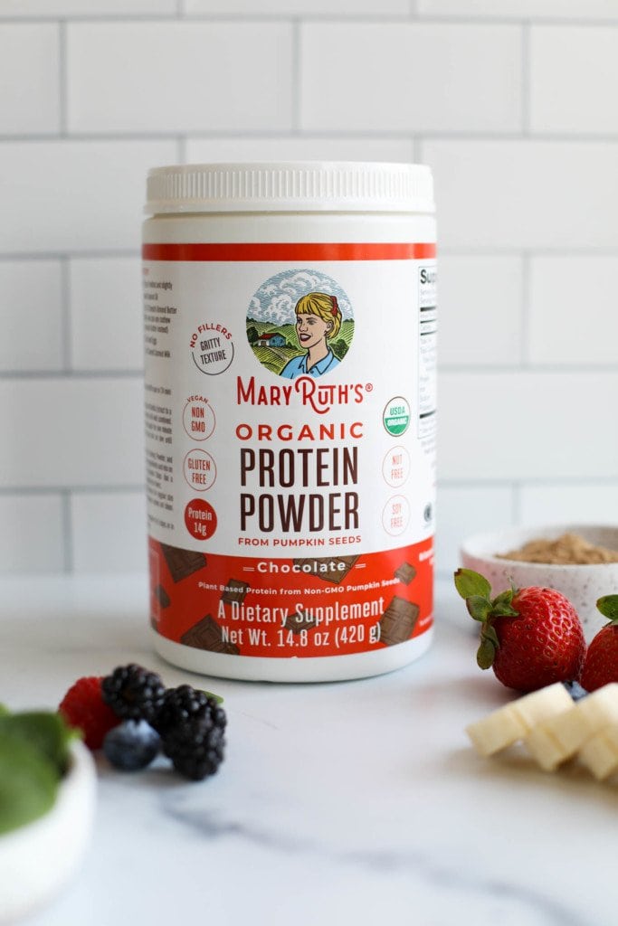 A container of Mary Ruth's Organic Plant Based Protein Powder with fresh fruit in the forefront. 