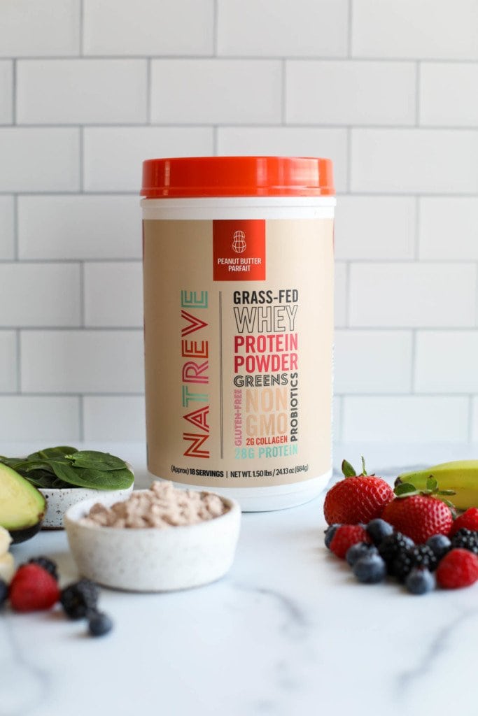 A container of Natreve grass-fed whey protein powder against a white background with fresh fruits and spinach in the forefront. 