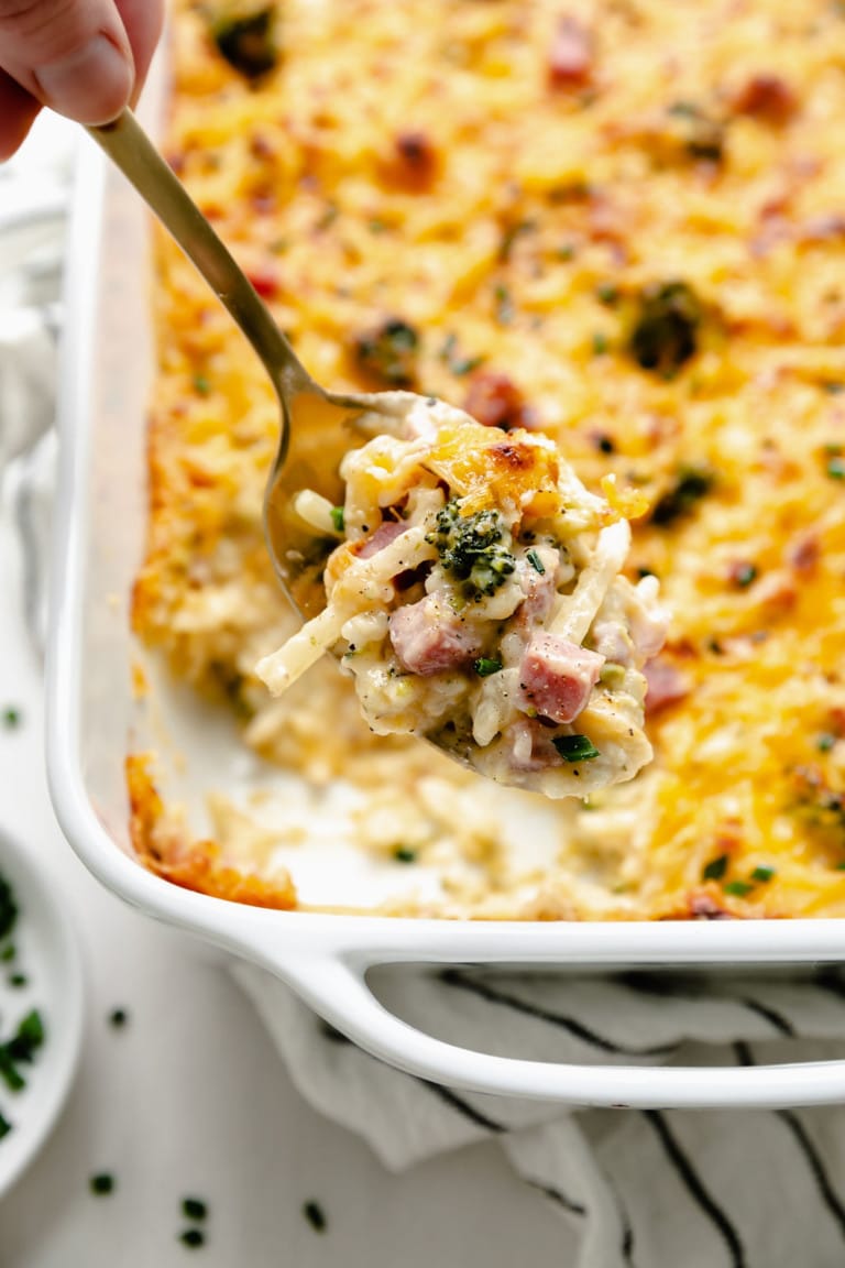 Cheesy scalloped potatoes and ham casserole with broccoli scooped up on a gold spoon over a white casserole dish.