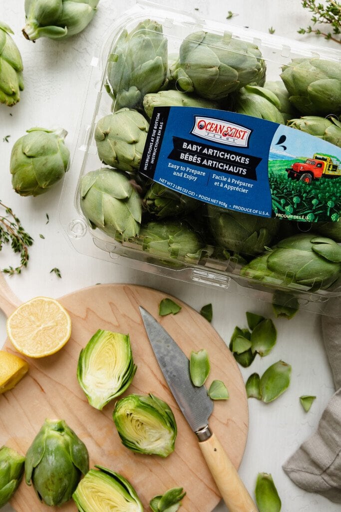 Overhead view of a package of Ocean Mist baby artichokes next to a light wooden cutting board with a baby artichoke cut in half to show inside texture. 