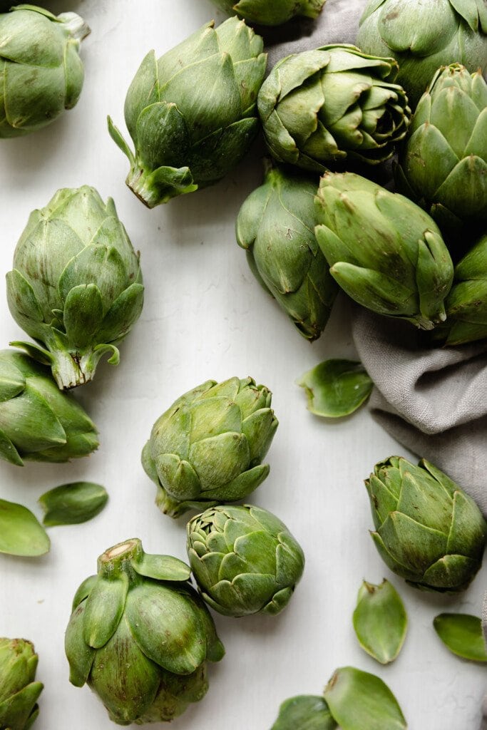 Several baby artichokes arranged together on a white background with a light tan dishcloth to one side. 