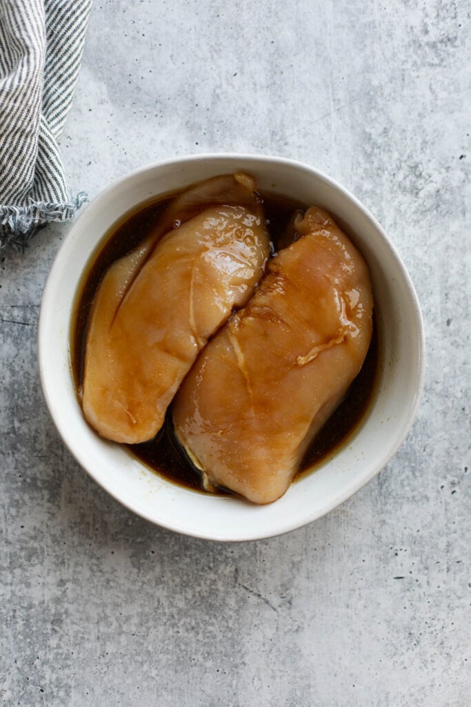 Overhead view of chicken breasts in teriyaki marinade in a white bowl. 