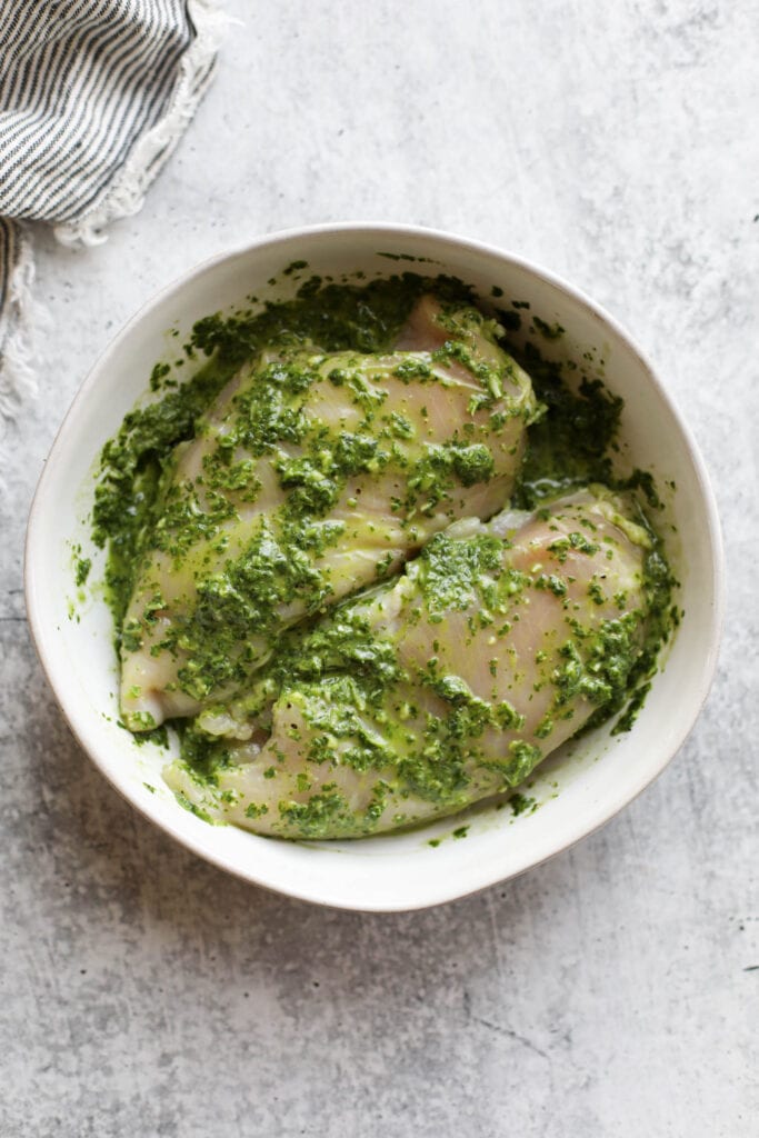 Overhead view of two chicken breasts in basil pesto chicken marinade in white bowl.