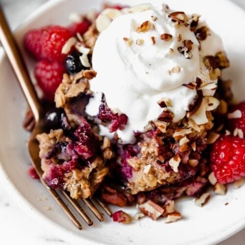 A piece of mixed berry baked oatmeal on a white plate topped with yogurt, nuts, and fresh raspberries.