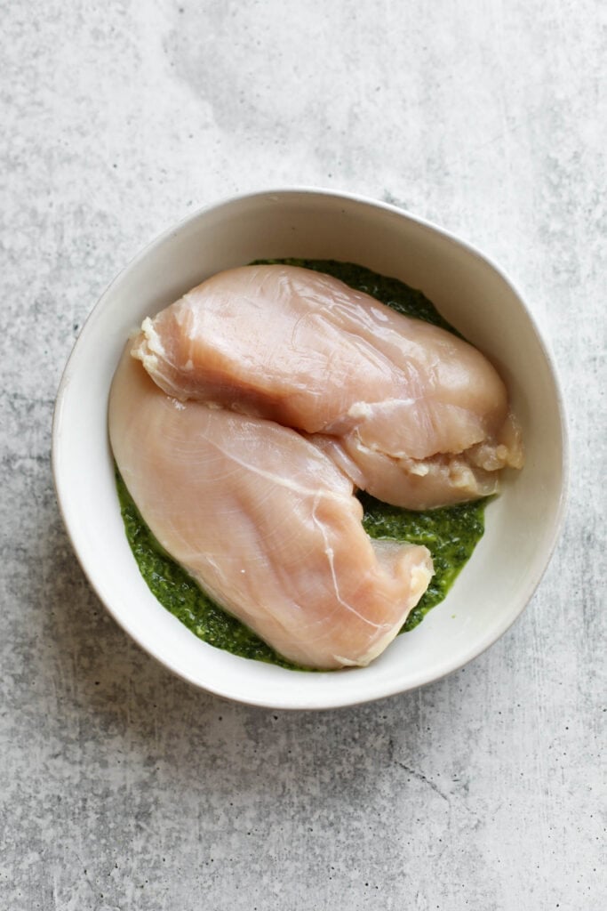 Overhead view of two chicken breasts in a white bowl with basil pesto chicken marinade. 