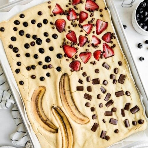 Overhead view of easy sheet pan pancakes in a baking sheet topped with blueberries, strawberries, banana slices, and chocolate chunks.