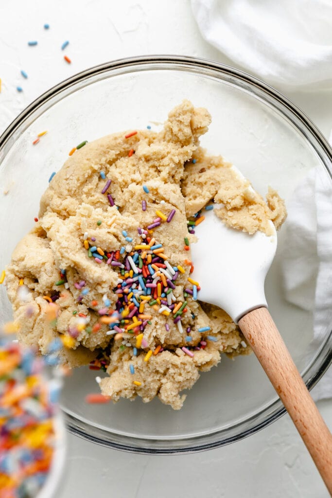 Funfetti cookie dough in a clear mixing bowl with a white spatula mixing the dough, a bowl of colorful sprinkles being poured into the dough.