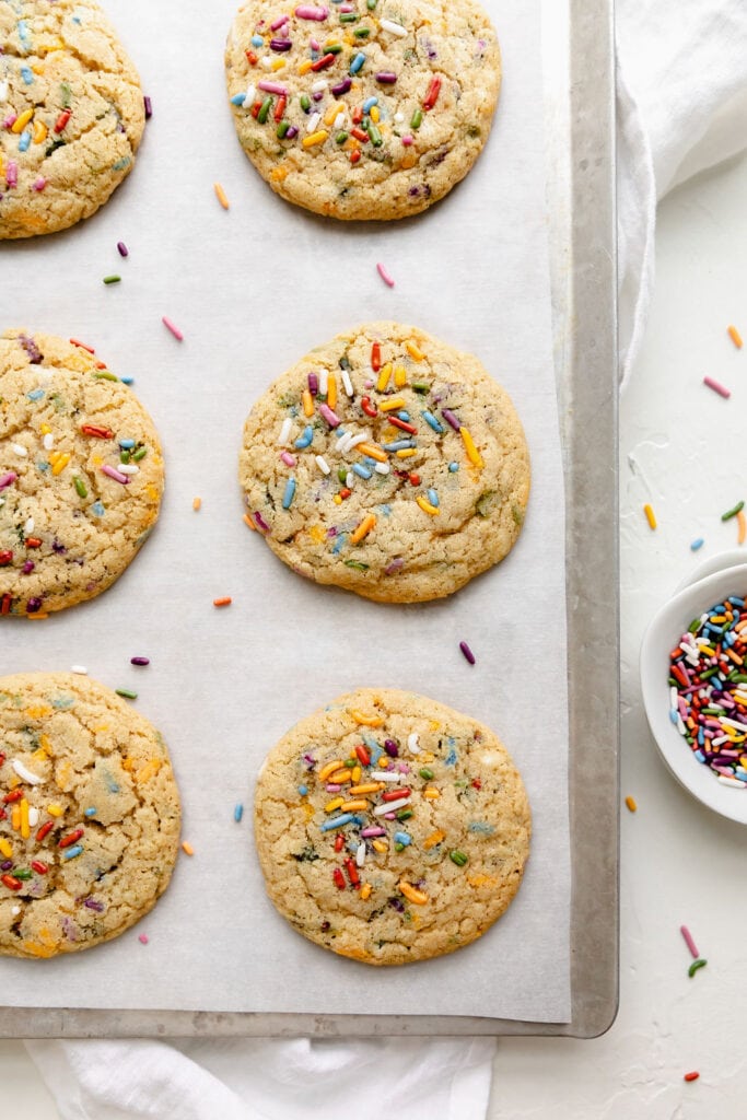 Overhead view of freshly baked gluten free funfetti cookies lined up on a parchment-lined baking sheet, extra colorful sprinkles on top each cookie. 