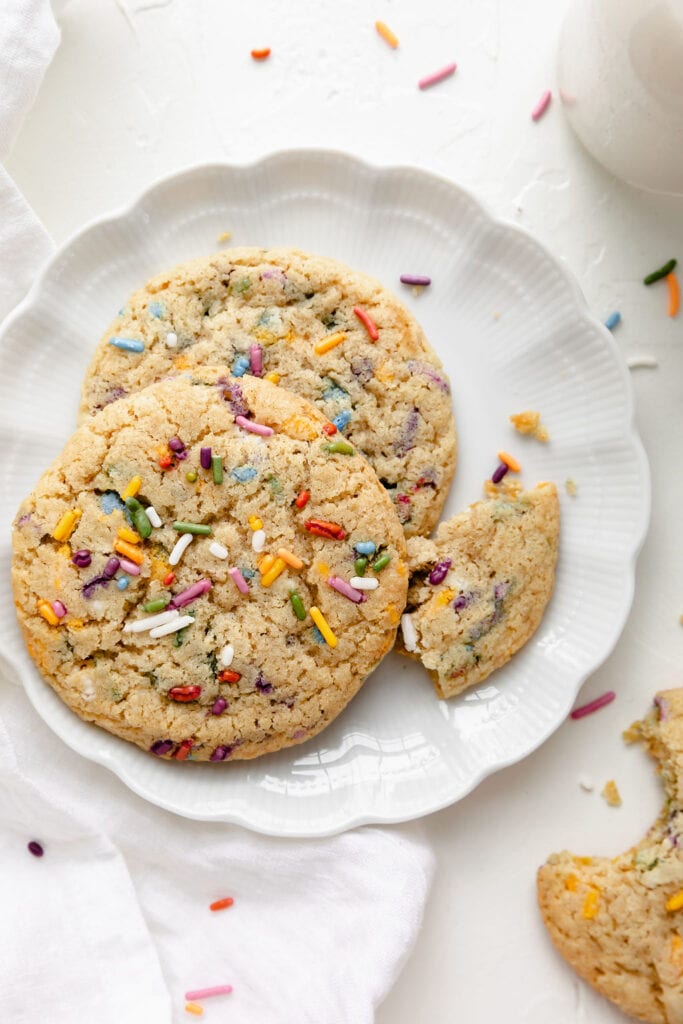 Two gluten free funfetti cookies on a scalloped edge white plate, a small morsel of a cookie on the edge of the plate from being eaten. 