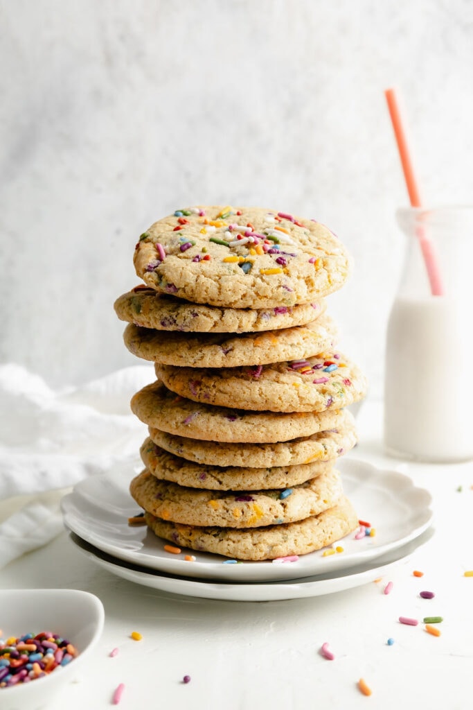 A tall stack of gluten free funfetti cookies on a scalloped edge white plate, a small glass jug of white milk with an orange straw in the background. 