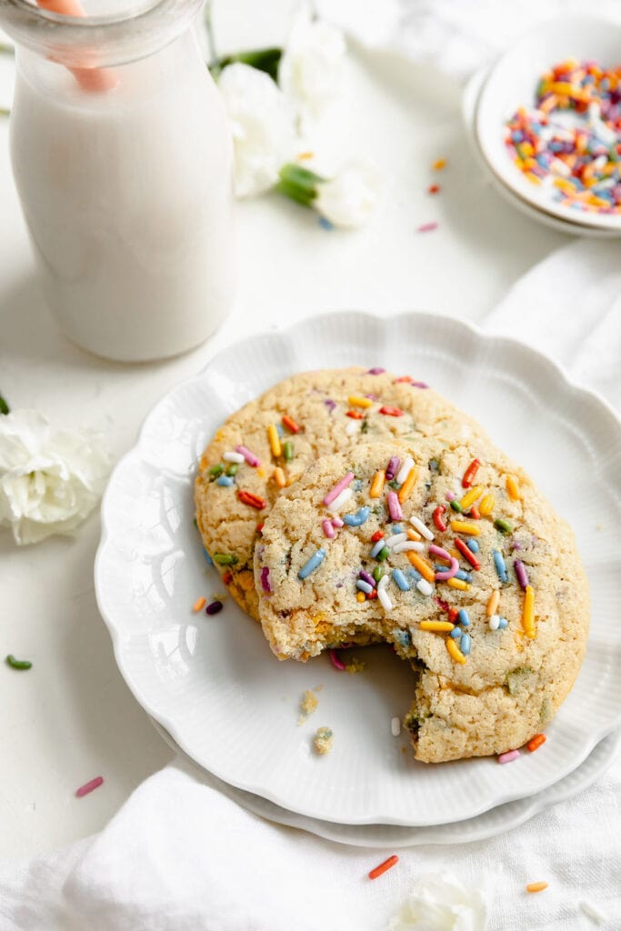 A scalloped edge white plate with two funfetti cookies, one with a bite taken out, on the plate, extra sprinkles on top each cookie. A jar of white milk with an orange straw in the background. 