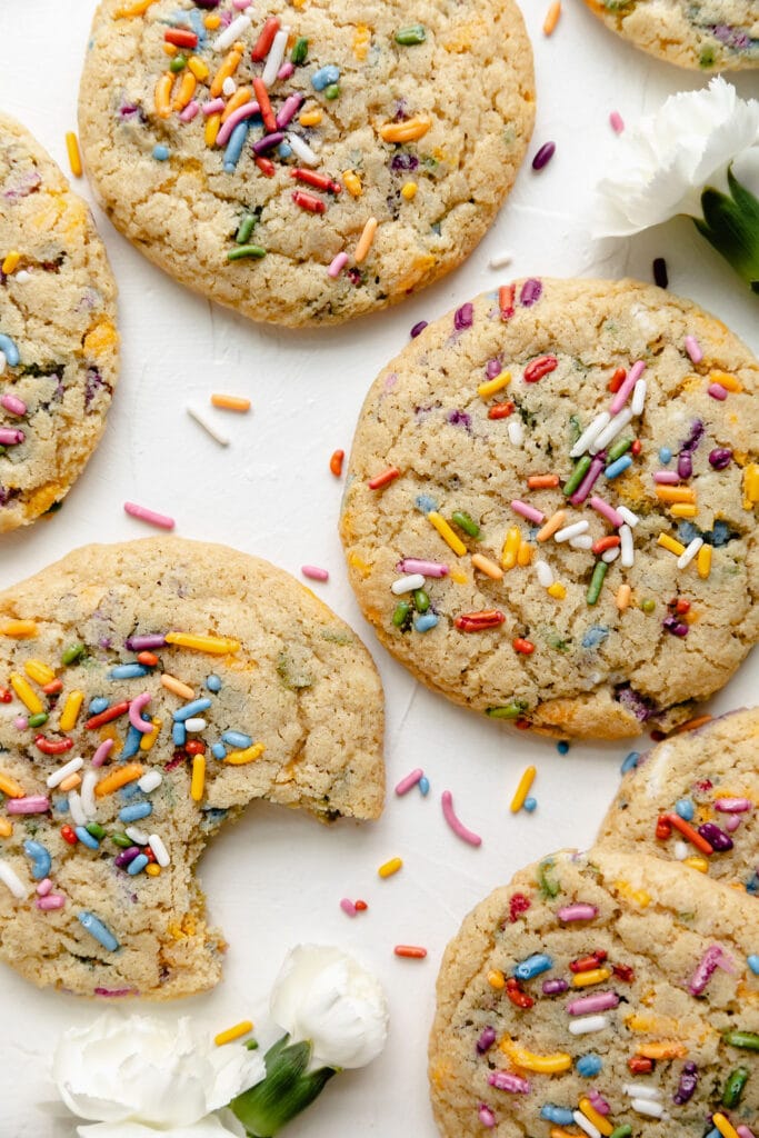 Overhead view of several funfetti cookies laying on a counter with extra colorful sprinkled scattered around, a couple small white flowers nearby, and a bite taken from one cookie.