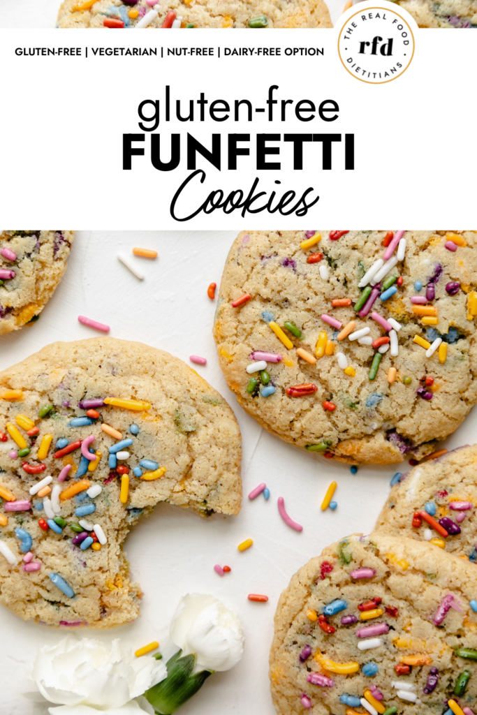 Overhead view of several gluten free funfetti cookies topped with extra colorful sprinkles arranged on a white counter with small white flowers in the corner. 