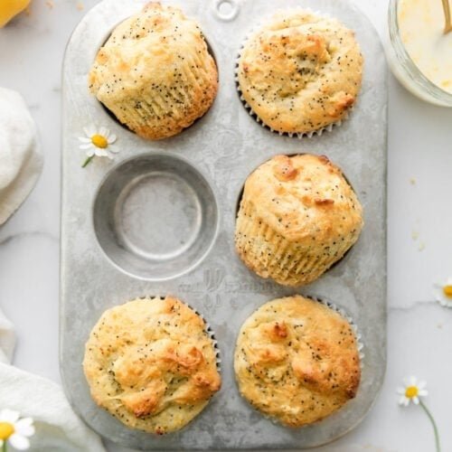 A silver six count muffin tin filled with five lemon poppyseed muffins, one muffin on the counter topped with lemon glaze.