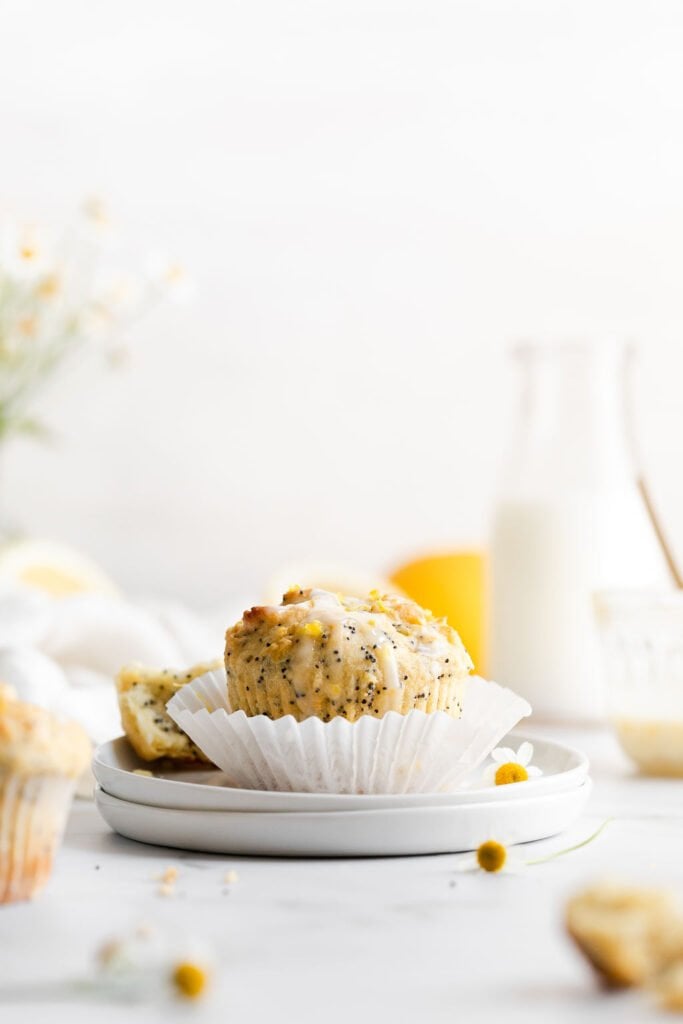 A single lemon poppy seed muffin topped with lemon glaze and sprinkled with lemon zest on a fanned out white muffin liner sitting on two stacked up white plate. 