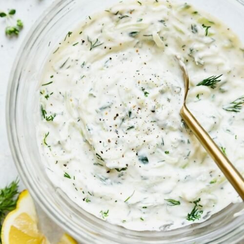 Overhead view of homemade tzatziki sauce in a clear glass mixing bowl with gold spoon in sauce.