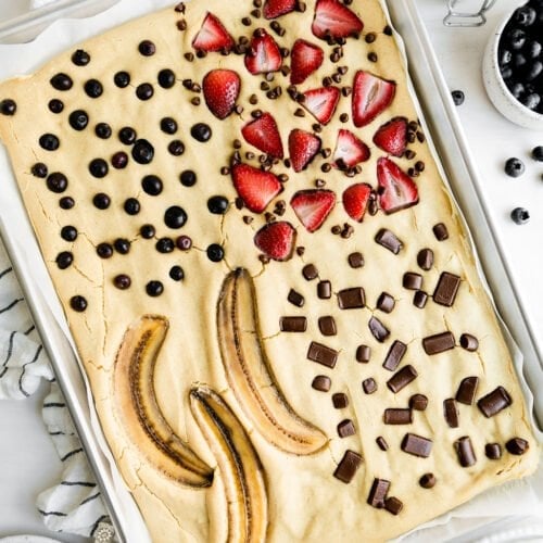 Overhead view of sheet pan pancakes baked in a silver baking sheet topped with strawberries, blueberries, chocolate chunks, and sliced bananas.