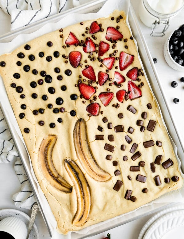 Overhead view of sheet pan pancakes baked in a silver baking sheet topped with strawberries, blueberries, chocolate chunks, and sliced bananas.