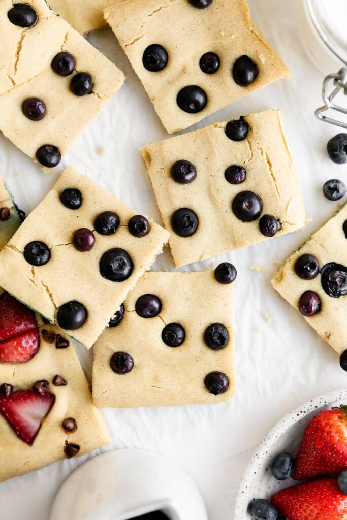 Several squares of sheet pan pancakes topped with blueberries arranged on a piece of parchment paper. 