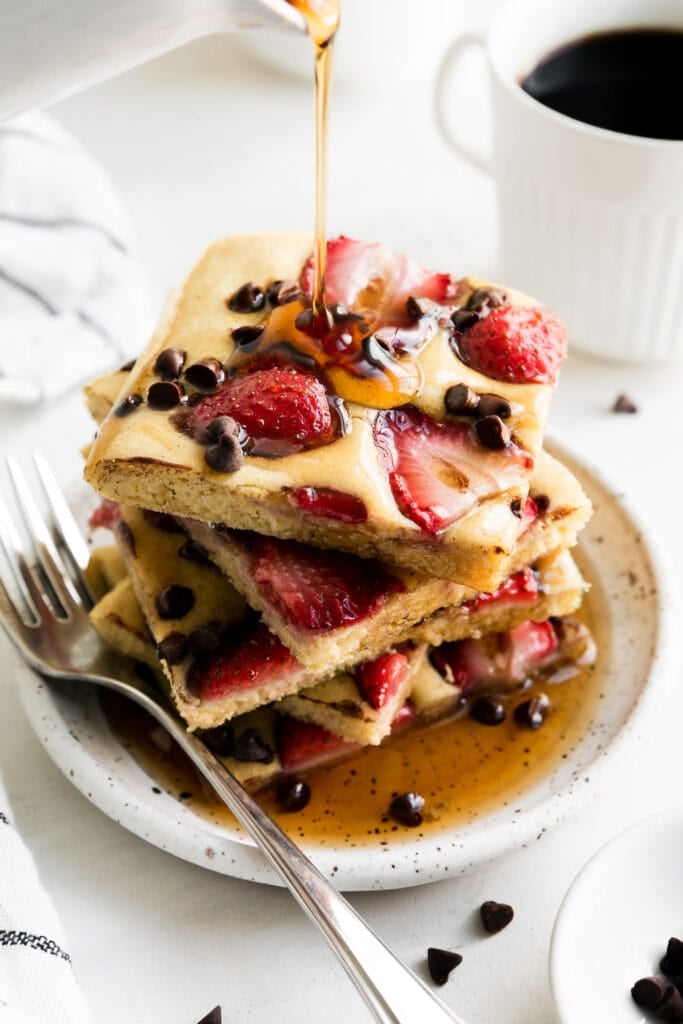 A stack of sheet pan pancakes cut into squares, stacked on top of each other, topped with strawberries and mini chocolate chips. 