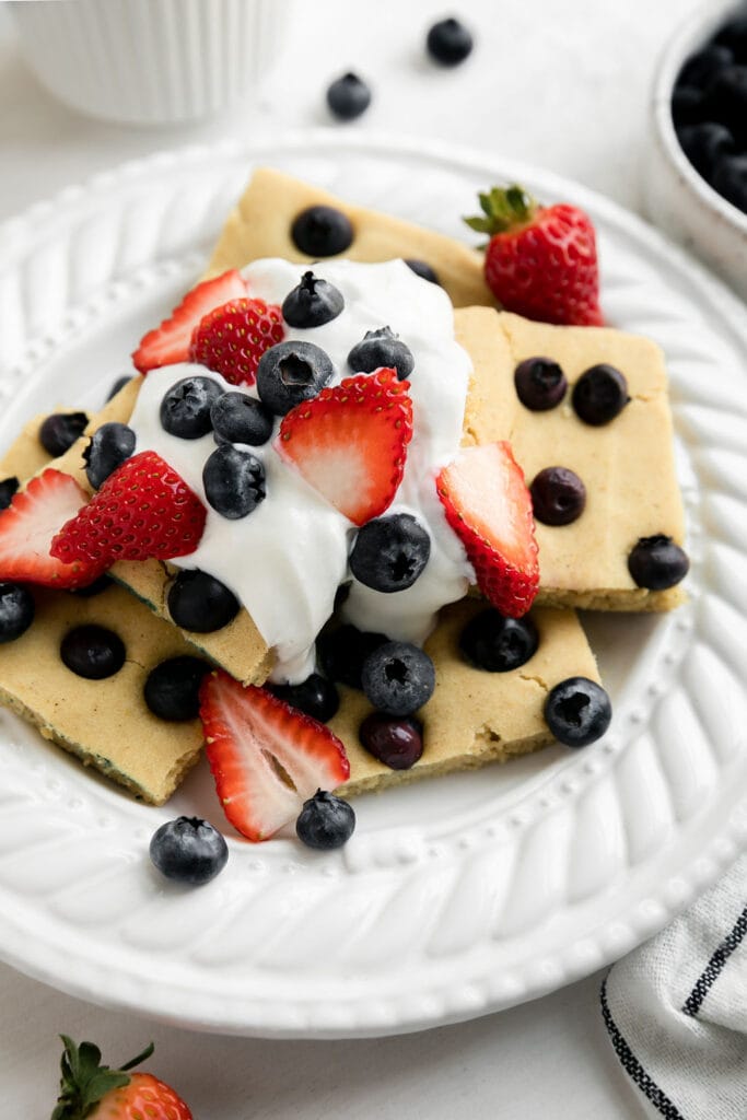 A pile of sheet pan pancakes cut into squares served on a white plate topped with whipped cream and fresh blueberries and sliced strawberries. 