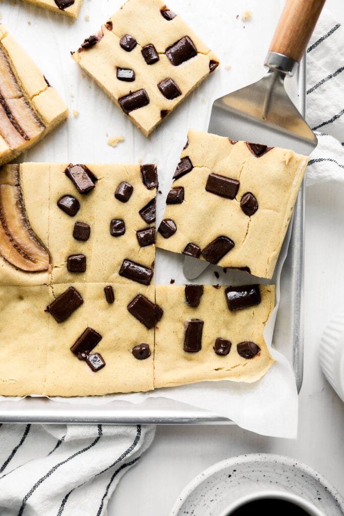 Sheet pan pancakes baked in a parchment lined baking sheet cut into squares, topped with chocolate chunks, with one square being lifted up with a cake server. 