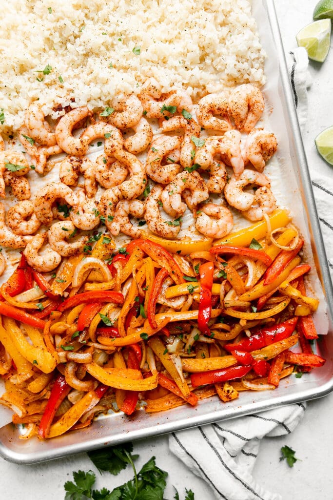 Overhead view of shrimp fajitas on a sheet pan; one-third pan filled with bell peppers and onions, one-third with seasoned shrimp, and one-third with cauliflower rice. 