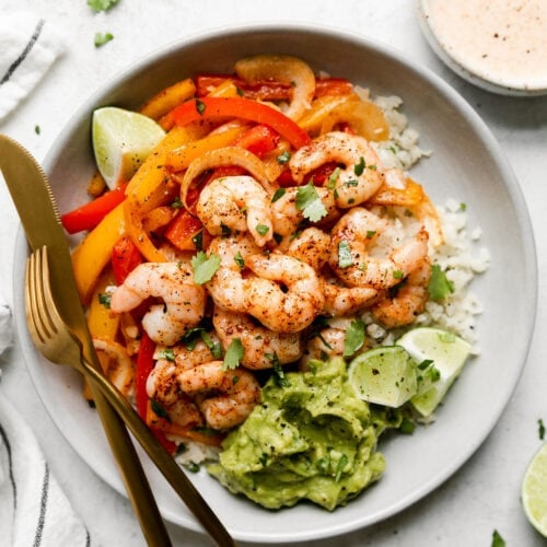 Overhead view of sheet pan shrimp fajita bowls served on a plate layered with cauliflower rice, peppers and onions, and topped with fajita shrimp. Avocado and limes on the side.