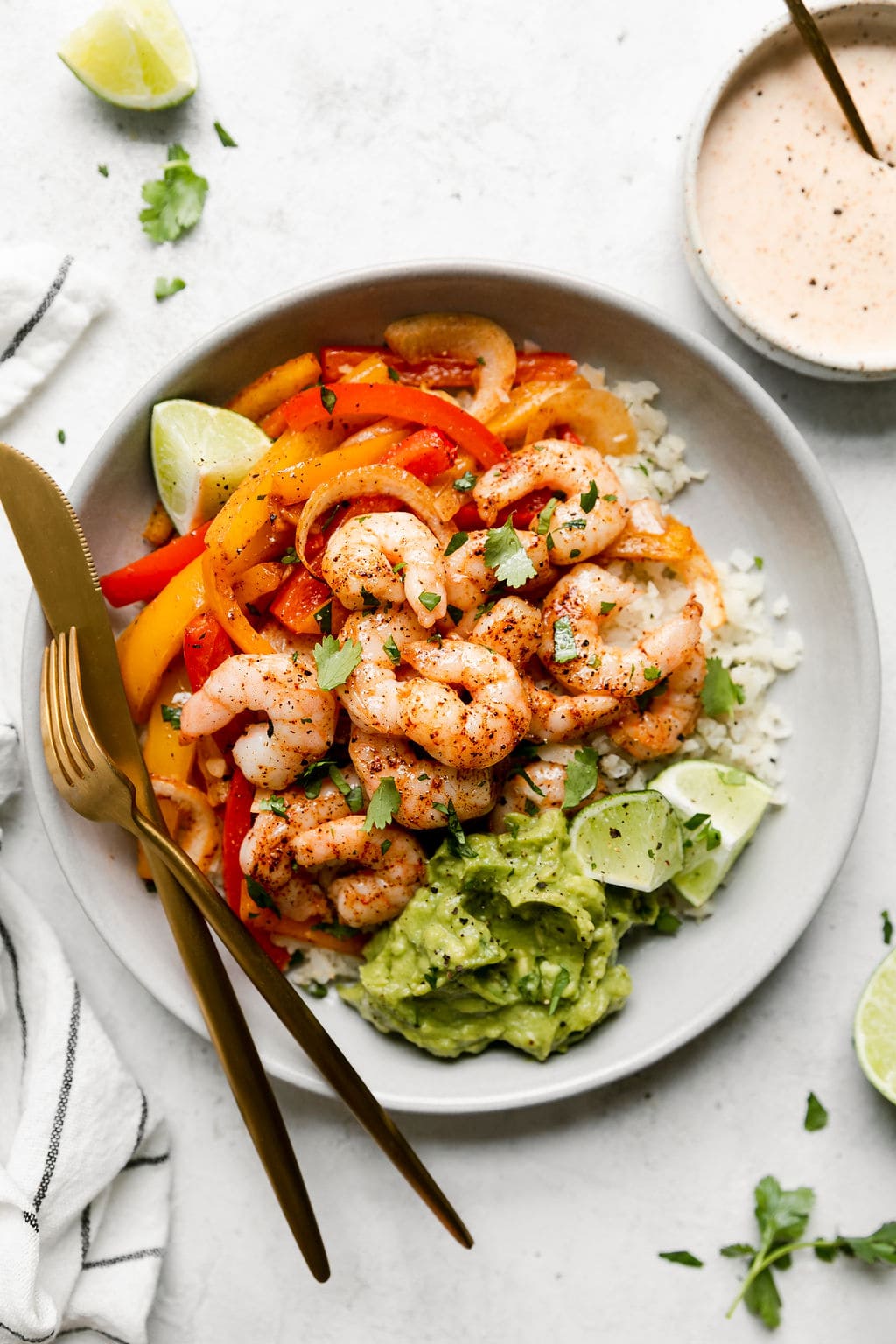 Overhead view of sheet pan shrimp fajita bowls served on a plate layered with cauliflower rice, peppers and onions, and topped with fajita shrimp. Avocado and limes on the side.