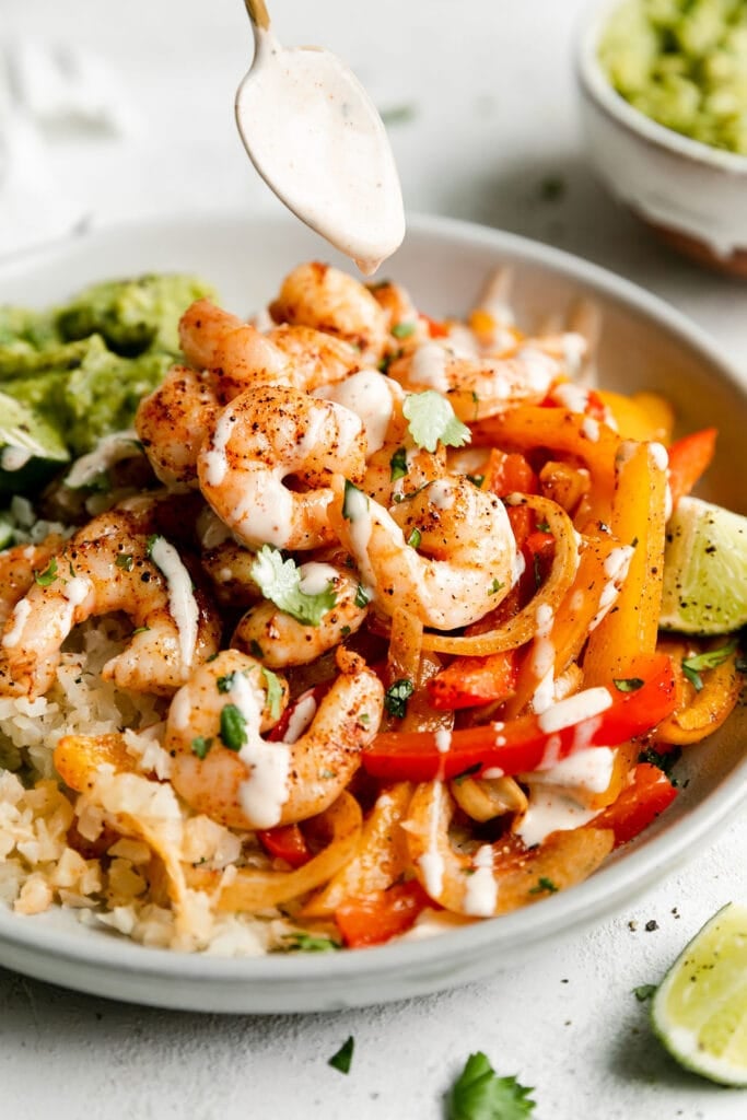 A spoon hovering over a bowl filled with sheet pan shrimp fajitas drizzling homemade ranch dressing over the pink shrimp.