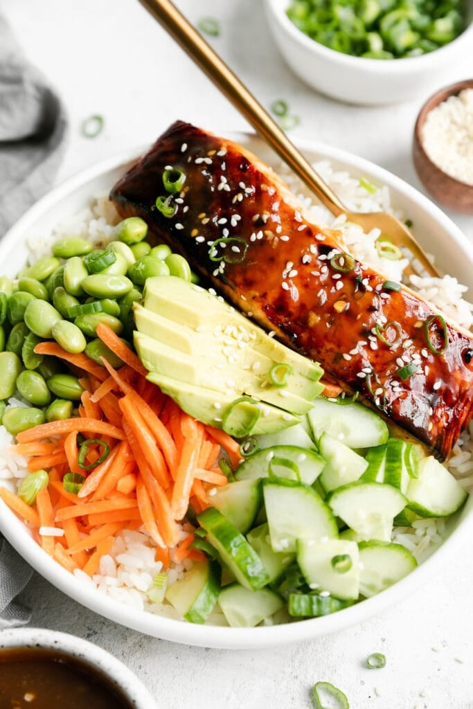 Teriyaki Salmon Bowls served in a white bowl over white rice topped with broiled salmon, edamame, carrots, avocado, cucumber and sesame seeds. 