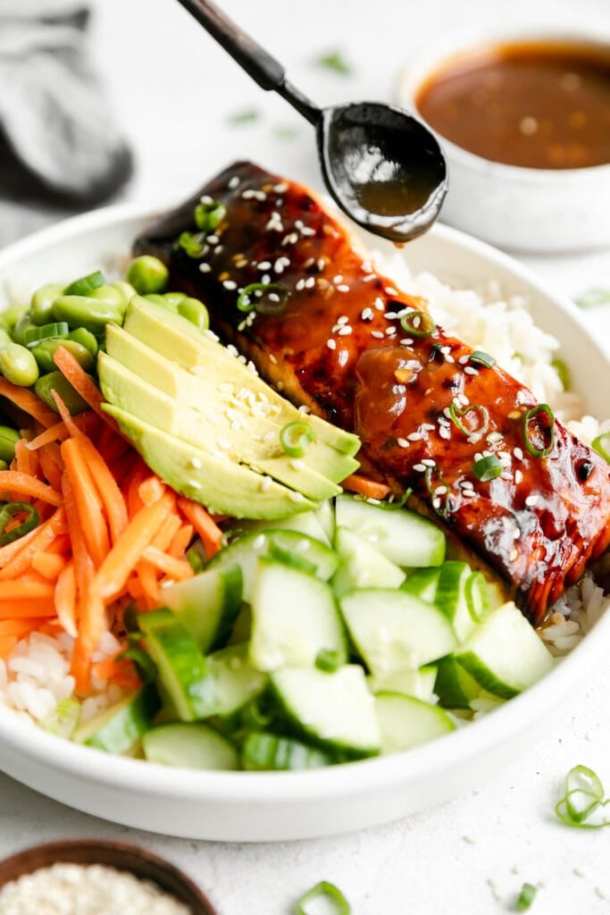Close up view of teriyaki salmon bowl served in a white bowl, topped with a broiled salmon filet with a spoon drizzling teriyaki sauce over salmon. 