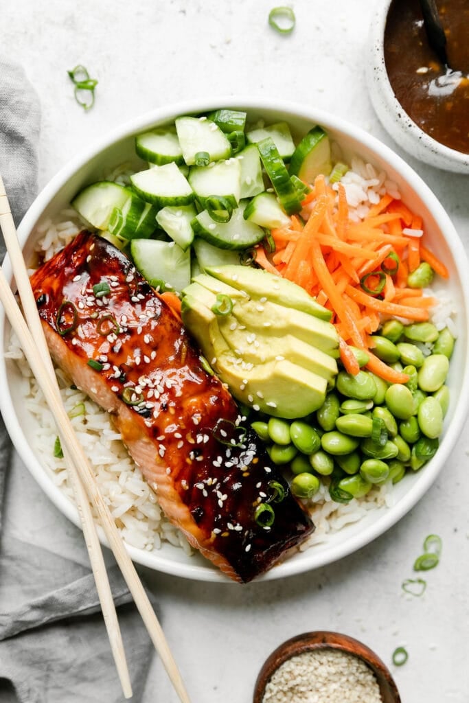 Overhead view of teriyaki salmon bowl served in a white bowl with rice, chopped cucumber, edamame, sliced avocado, and shredded carrots, chopsticks on the side of the bowl. 