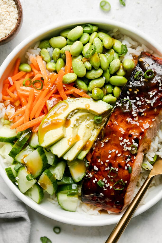 Close up overhead view of teriyaki salmon bowl served in a white bowl with rice, edamame, carrots, avocado, cucumber and broiled teriyaki salmon filet. 