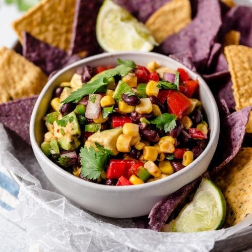 Black bean and corn salad in a white bowl with yellow and blue corn chips surrounding it.