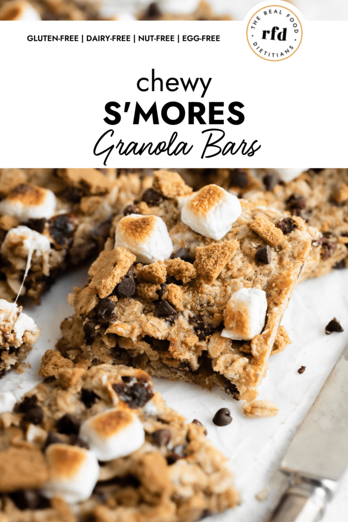 Close up view of a square cut chewy s'mores granola bar topped with toasted mini marshmallows and mini chocolate chips perched on more square granola bars. 