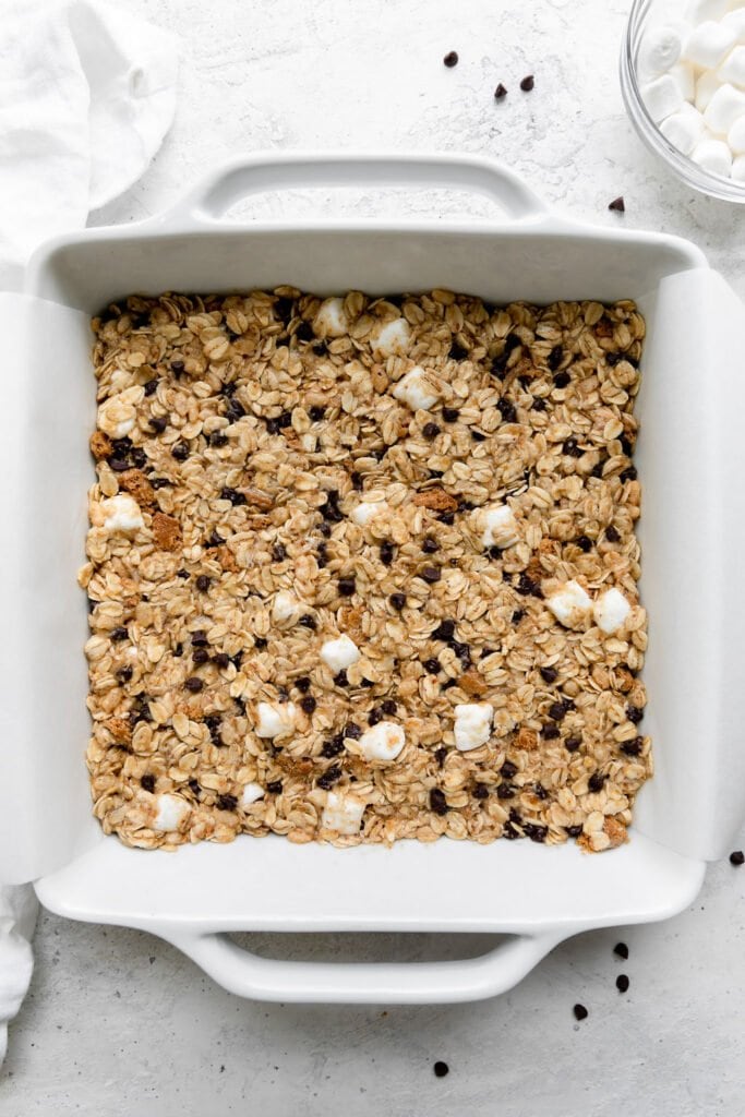 Overhead view of chewy s'mores granola bars pressed into 8x8 white baking dish ready for baking.