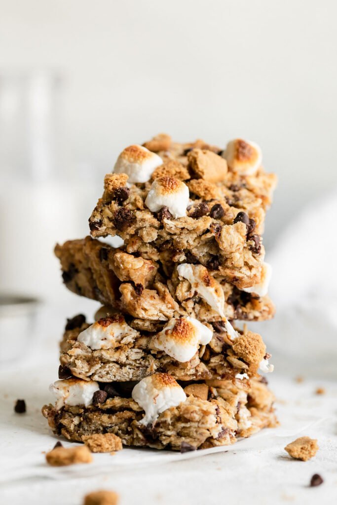 Four chewy s'mores granola bar squares stacked up on each other with toasted mini marshmallows and melty chocolate chips on top.