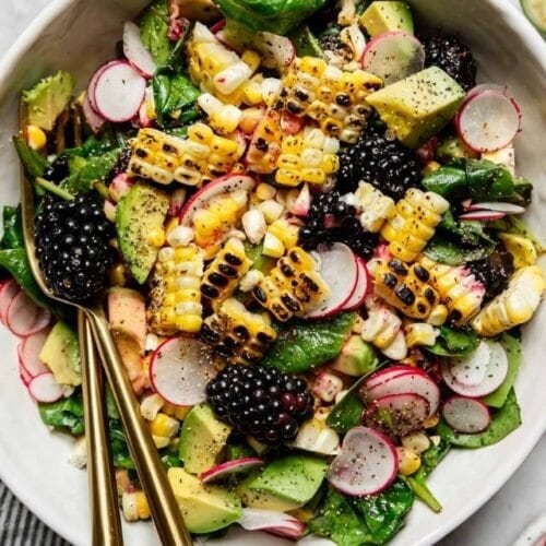 Overhead view grilled corn salad with blackberries in white bowl with gold cutlery.