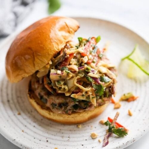 Grilled pork burger with creamy Thai slaw on toasted bun served on white speckled plate.